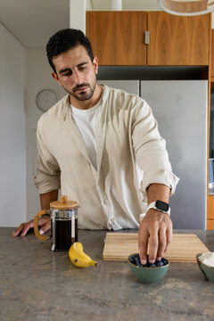 Adult man wearing beige shirt and smartwatch at kitchen island reaching into bowl of berries