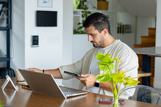 Adult man in 30s wearing light overshirt, sitting at home workspace checking smartphone and laptop