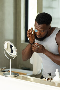 Adult male applying under-eye patch while checking mirror at vanity, wearing grey tank and bracelet