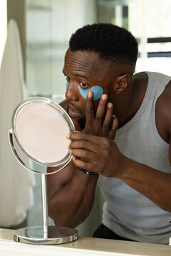African American man holding round mirror while applying blue eye patch at bathroom counter