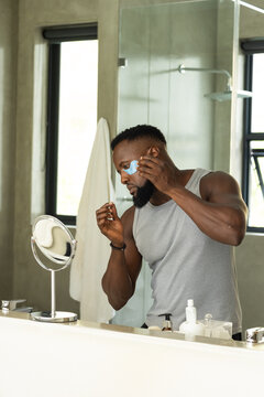 Man wearing grey tank and beaded bracelet standing at bathroom mirror applying blue under-eye patch