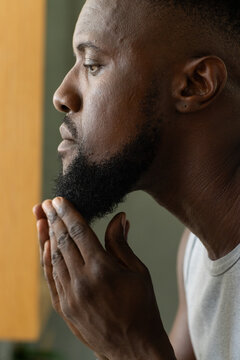 Man grooming beard at window with hands, wearing white sleeveless shirt and stud earring