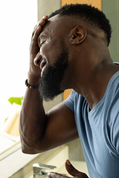 African man holding forehead while leaning at bathroom sink with chrome faucet and beaded bracelet