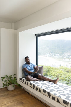Smartphone buzzing, African male checking wristwatch for time while sitting on window bench at home