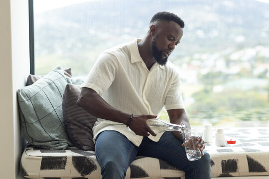 Adult man seeing white pill bottles, pouring water into glass on window bench, taking pills