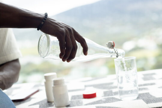 Man pouring water from swing-top bottle into tumbler at table by window, med bottles, bracelet