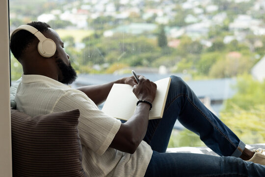 African man reclining on window seat wearing white headphones and light shirt, writing in notebook