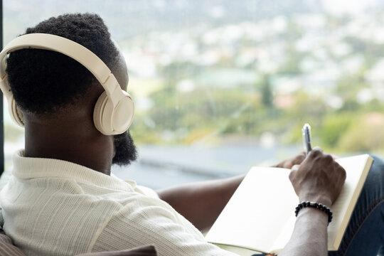 Adult African American man sitting by window wearing white tee headset writing notes, copy space