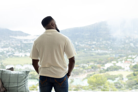 Adult African man wearing cream polo jeans standing at home noticing rain on glass, watching valley