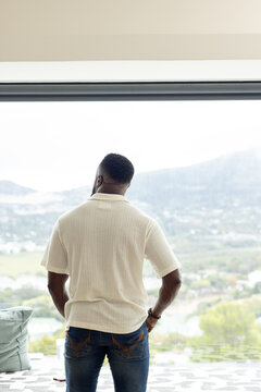 African American man standing by window in modern room wearing polo and jeans smartphone nearby