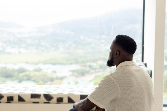 Adult African man sitting on patterned window seat in white polo gazing through glass, copy space