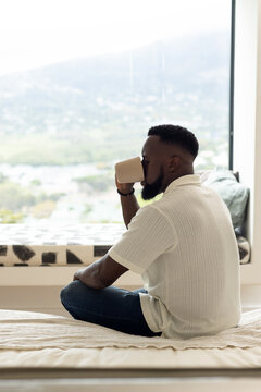 Adult African man sitting on bed by large window drinking from mug wearing beads, copy space