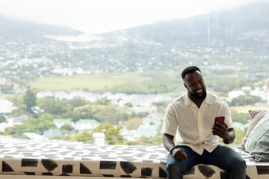 African man sitting on patterned bench by window holding red smartphone smiling, copy space