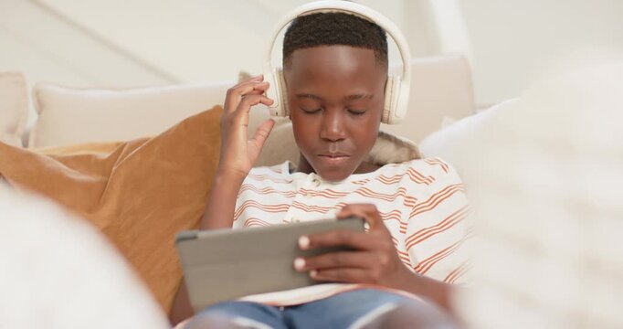 African American teen raising hand, donning headset, tapping tablet on sofa, focusing on content