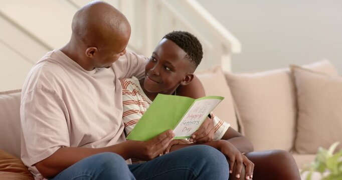 African father and teenage son opening green Father's Day card on couch, celebrating and hugging