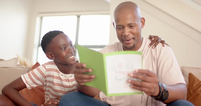 African dad, teen son after giving green handmade card on couch, dad reading, hugging, showing love