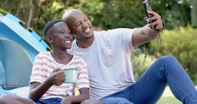 African American dad showing phone, teen son posing, both capturing moment on picnic blanket