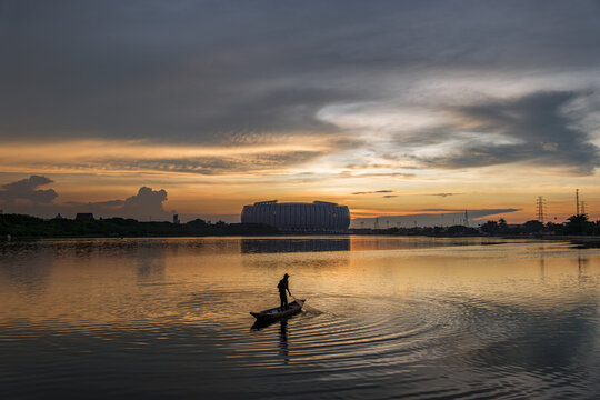 Striking contrast of a traditional fisherman on a boat at Danau Sunter lake during golden hour, with the modern futuristic architecture of Jakarta International Stadium (JIS) in the background, Indone