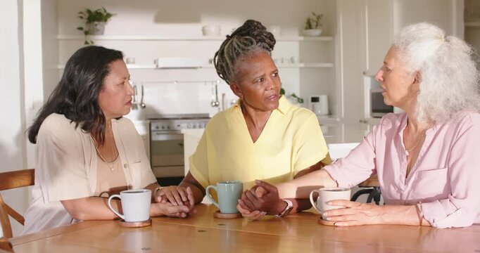 Diverse women holding hands at kitchen table, center turning, speaking, comforting with mugs, watch