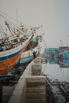 a row of traditional wooden cargo vessels, known as Pinisi or Lambo ships, docked along a concrete pier in what appears to be an Indonesian harbor,  Sunda Kelapa in Jakarta.