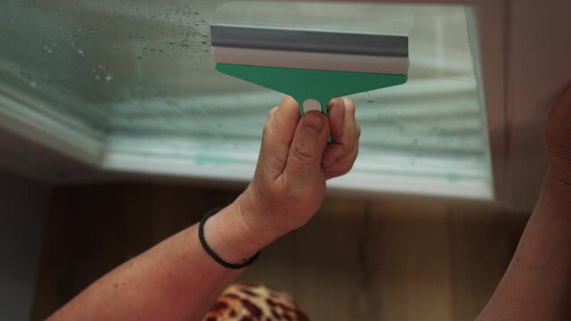 A person cleans a window with a green squeegee in a kitchen