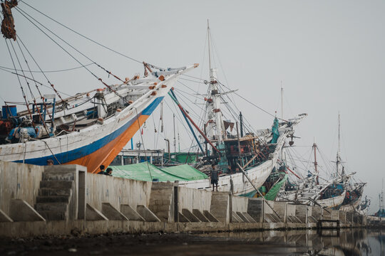 a sweeping view of the concrete pier at Sunda Kelapa harbor, dominated by the massive, angled hulls of traditional Pinisi ships.