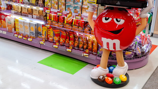 A giant red MM character mascot candy dispenser standing next to snack shelves in a modern supermarket aisle on March 28, 2026 in Bangkok, Thailand