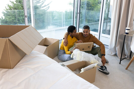 Unpacking boxes, Diverse couple sitting in bedroom near windows, examining bubblewrap picture frame