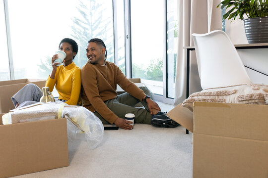 Holding coffee cups, diverse couple sitting on carpet by sliding glass doors, with moving boxes