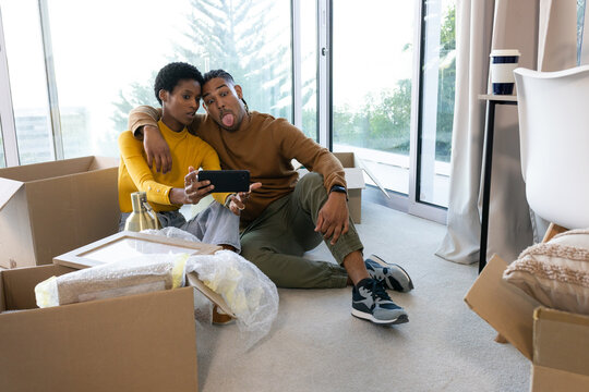 Smiling couple taking selfie on carpeted floor in new apartment, with boxes and bubble wrap