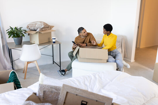 Sorting Diverse couple unpacking bubble wrap on ottoman in bedroom, with cardboard boxes