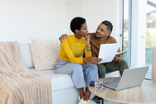 Diverse couple sitting on white sofa in living room studying documents, with open laptop and folder