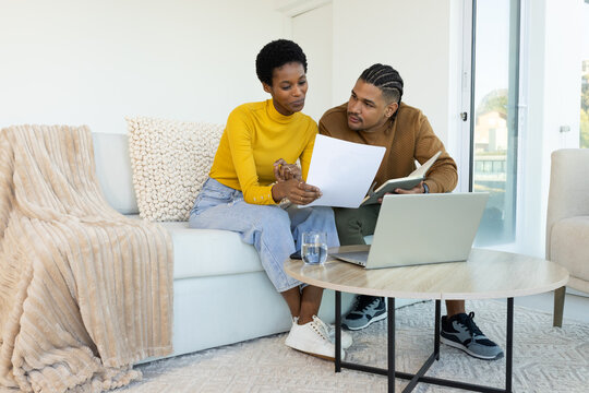 Diverse couple looking at documents and notebook on sofa in living room, with laptop, water
