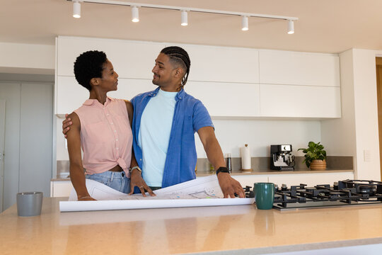Diverse couple reviewing rolled blueprints at modern kitchen island, with coffee maker and plant