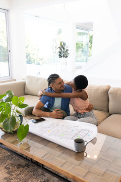 Hugging diverse couple on sectional couch in bright living room, with architectural blueprint