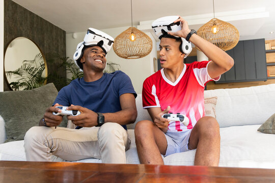Male friends gaming on light-gray sofa in living room, using VR headsets and controllers