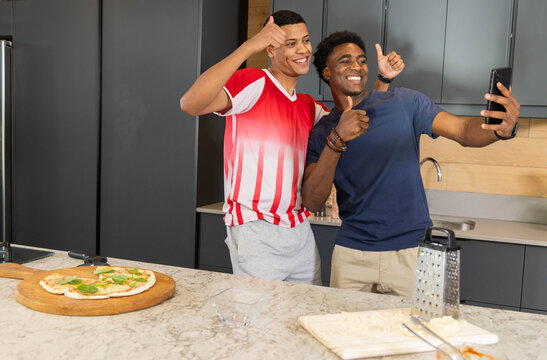 Taking selfie diverse male friends standing at modern kitchen countertop, with smartphone and pizza