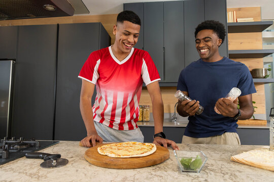 Smiling Diverse male friends preparing pizza on marble island in modern kitchen with pizza cutter
