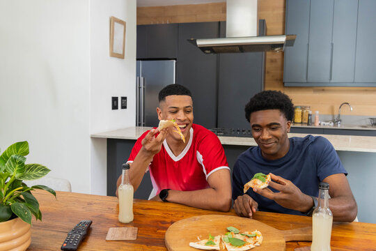 Diverse male friends sitting at kitchen table sharing pizza slices on pizza board with beverages