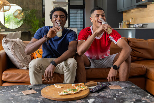 Diverse male friends drinking bottles on leather sofa in living room, with pizza on marble table