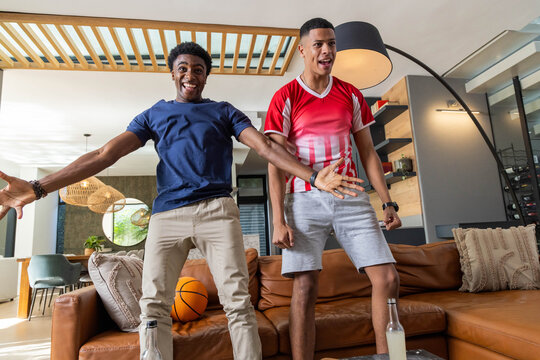 Standing diverse male friends spreading arms on sofa in modern living room, with basketball