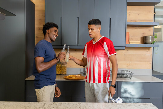 Clinking glass bottles diverse male friends wearing sportswear at kitchen island, with pizza slices