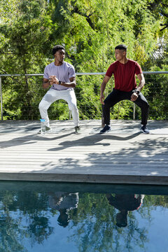 Squatting Diverse male friends in workout clothes on wooden deck next to pool, with water bottle