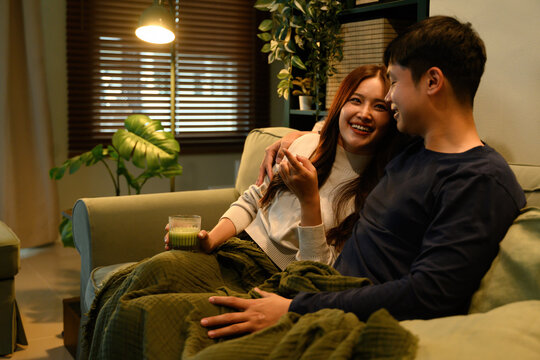 Asian couple relaxing on a sofa, watching TV together with smiles, enjoying a cozy and intimate evening at home.
