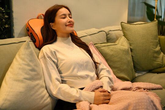 Asian woman relaxing on a sofa using an ergonomic neck massage pillow, enjoying comfort and stress relief in a cozy home