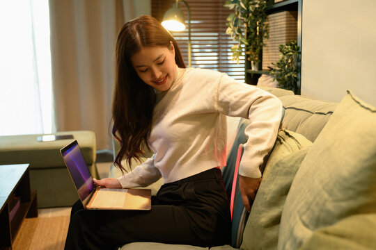 An Asian woman adjusting a back massage device while sitting on a sofa with a laptop, promoting ergonomic support and work-from-home wellness