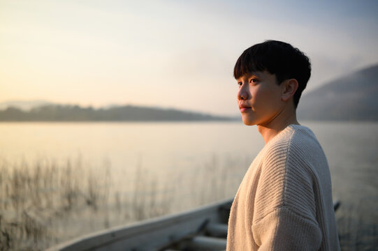 Close portrait of a young Asian man standing by a peaceful lake at sunrise, looking into the distance with warm sunlight on his face, expressing quiet confidence and reflective mood