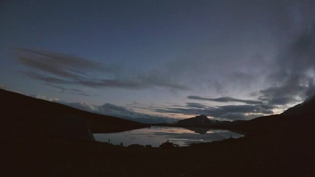 Day to night timelapse Magical twilight at Koruldi Lakes, Georgia. A glowing yellow tent sits by the mirror-like water, reflecting the vast Caucasus peaks under a starry mountain sky