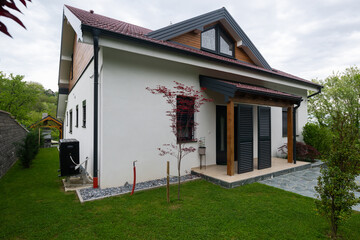 Fototapeta premium Modern white suburban house with covered porch, dark shutters and landscaped lawn under overcast sky