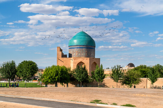 Turkistan, Kazakhstan - June 2, 2025: Scenic exterior view of the Rabia Sultan Begum Mausoleum with its turquoise dome and a flock of birds flying in the blue sky